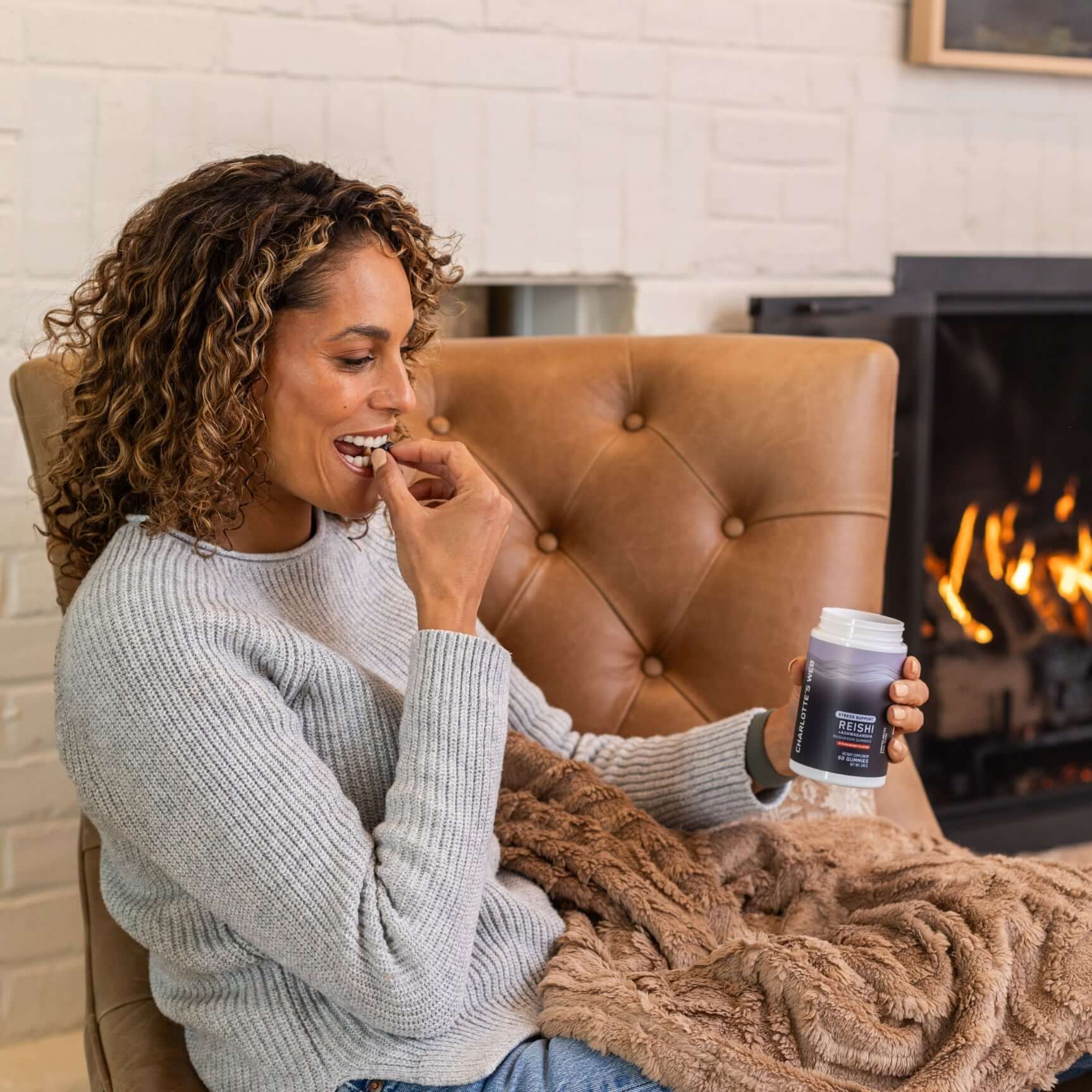 Women sitting in a brown chair by a fireplaece  holding a jar of STRESS SUPPORT WITH REISHI & ASHWAGANDHA MUSHROOM GUMMY - Strawberry flaovr while putting a gummy in her mouth.