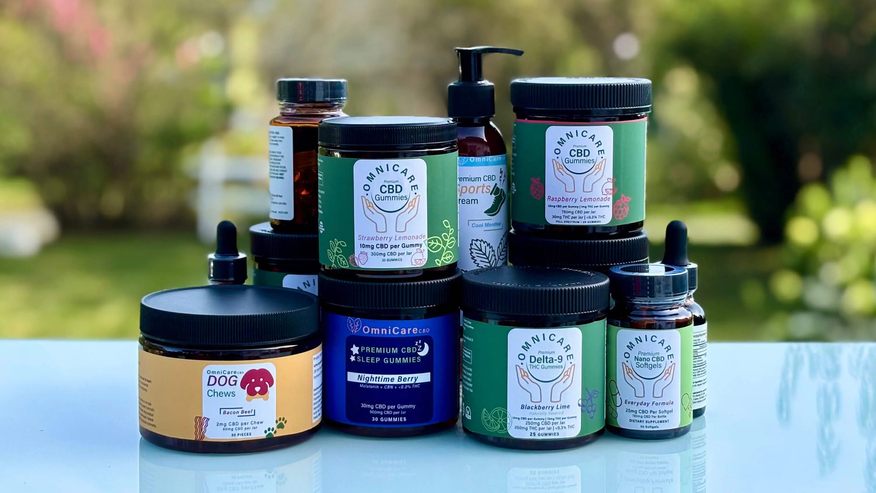 Collection of OmniCare CBD Delta 9 THC & CBD products — gummies, oils, tinctures, softgels, creams, topicals, and pet chews — displayed on a reflective surface with a blurred natural background.